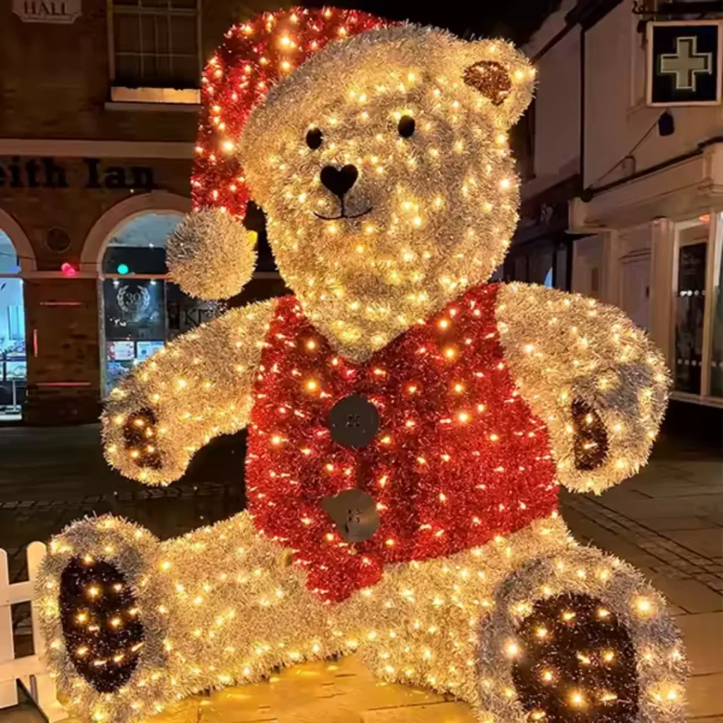 A giant illuminated teddy bear wearing a Santa hat and red vest, glowing against a festive town backdrop.