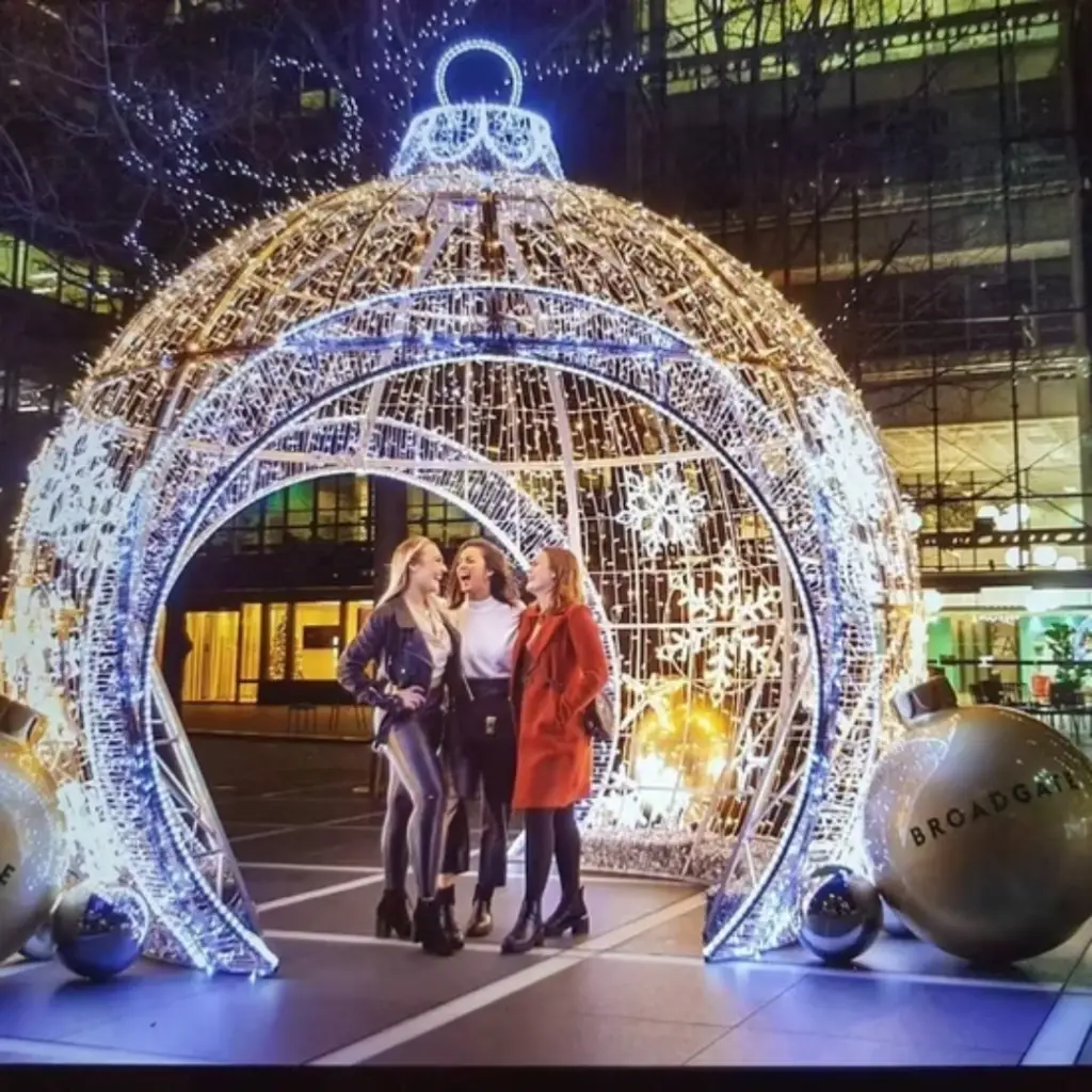 A grand illuminated Christmas ornament installation with an arched walkway, adorned with twinkling white and gold lights. Three women stand inside, laughing and enjoying the festive atmosphere. Large decorative ornament props labeled "BROADGATE" sit beside the structure, adding to the holiday charm.