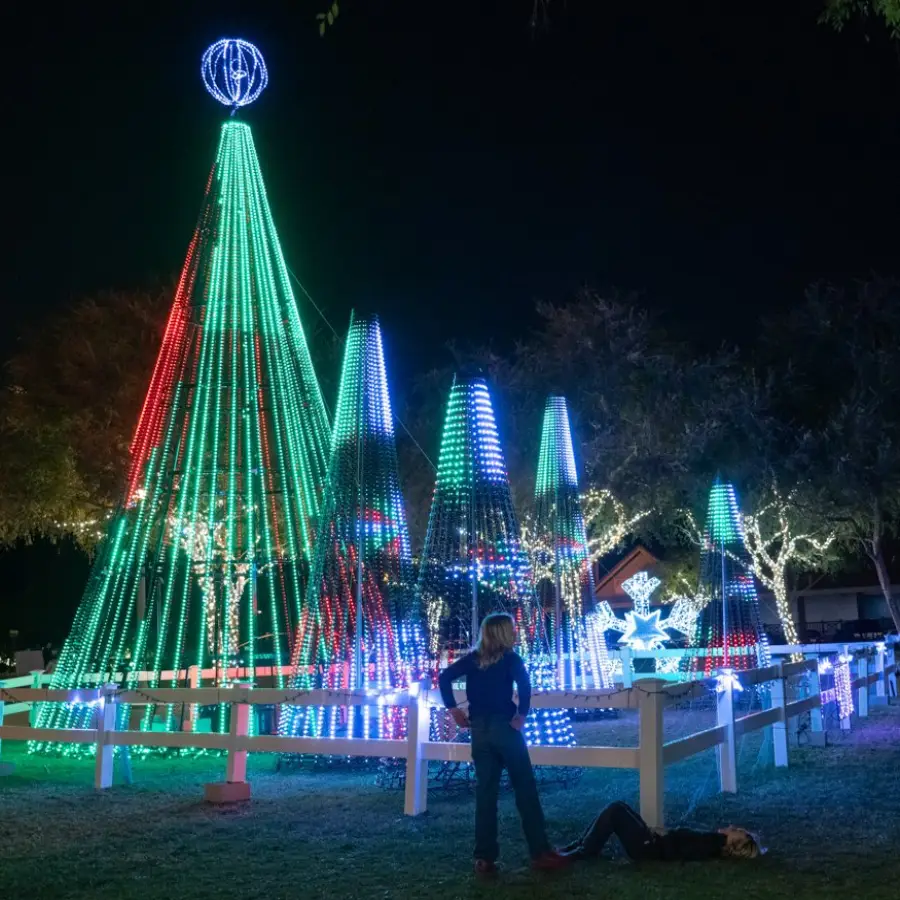 A dazzling Christmas light display featuring towering cone-shaped LED trees in red, green, and blue patterns. A glowing sphere sits atop the tallest tree, while additional illuminated decorations, including snowflakes and wrapped trees, enhance the festive scene. Two people, one standing and one lying on the ground, admire the display behind a white fence.